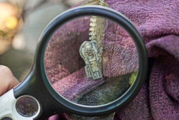 Fototapeta na wymiar a black magnifying glass in a hand magnifies a gray worn metal zip on a pink dirty jacket