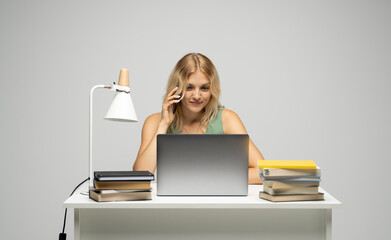 Cheerful blonde freelancer woman in green t-shirt talks on mobile phone with a clients while working on laptop in modern light office studio. Corporate Communication.