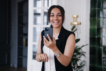 Fototapeta premium Happy young African American woman in whit shirt and black jacket holding phone looking at camera with wide smile at office, satisfied by new job. Successful Brazilian girl received awesome message.