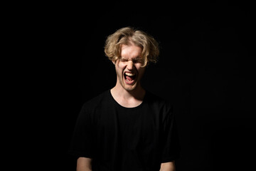 Angry, sad, aggressive young man in a black t-shirt screaming on a black background. Emotional...