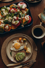 fried egg with avocado sandwich on a plate and caprese salad with colorful tomatoes and basil leaves. serving breakfast with black coffee top view.	