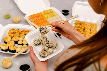 Woman eating fish sushi rolls with salmon,sesame,ginger,wasabi,soy sauce packed in plastic disposable container box on gray background.Seafood,Food delivery,take out service from restaurant concept