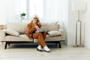 a cheerful stylish elderly woman is sitting in a brown suit with white boots and working at a laptop holding it on her lap while in a cozy apartment
