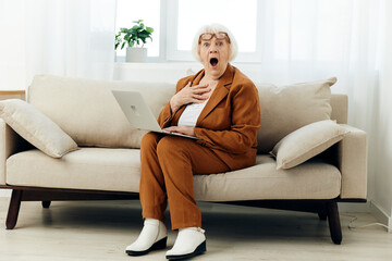 a shocked elderly woman with gray hair is sitting on a beige sofa in a brown suit working at a laptop and looking very emotionally amazed with surprise