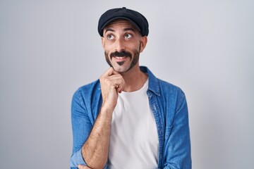 Hispanic man with beard standing over isolated background with hand on chin thinking about question, pensive expression. smiling and thoughtful face. doubt concept.