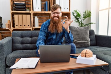 Redhead man with long beard using laptop working at psychology clinic doing ok sign with fingers,...