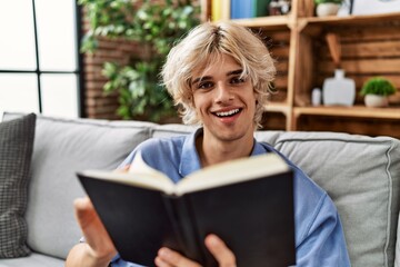 Young blond man reading book sitting on sofa at home