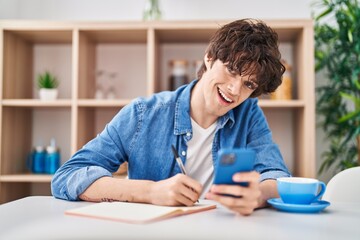 Young hispanic man write on notebook using smartphone at home