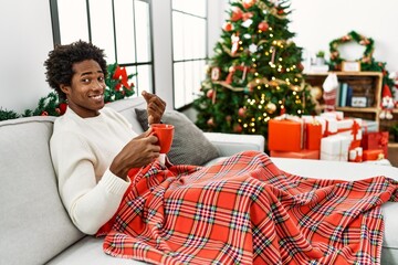 Young african american man sitting on the sofa drinking coffee by christmas tree doing money...