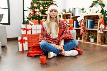 Obraz na płótnie Canvas Beautiful blonde woman sitting on the floor by christmas tree with serious expression on face. simple and natural looking at the camera.
