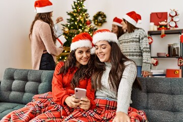 Group of young hispanic women on christmas meeting. Two woman sitting on the sofa using smartphone at home.