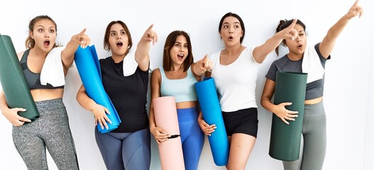 Group of women holding yoga mat standing over isolated background pointing with finger surprised...