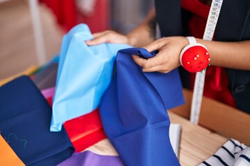 Young beautiful hispanic woman tailor holding cloths at clothing factory