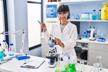 Young hispanic woman working at scientist laboratory smiling happy pointing with hand and finger to the side