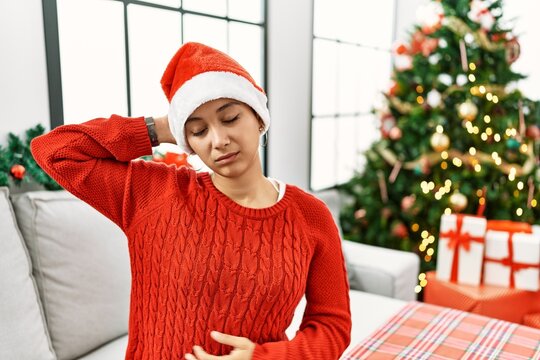 Young hispanic woman with short hair wearing christmas hat sitting on the sofa suffering of neck ache injury, touching neck with hand, muscular pain