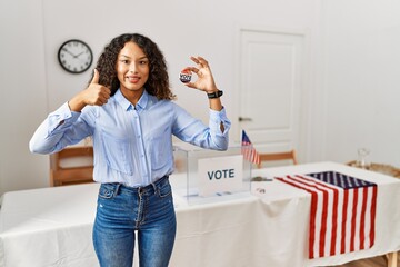 Beautiful hispanic woman standing by at political campaign by voting ballot doing happy thumbs up...