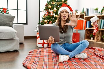 Young latin woman using laptop sitting by christmas tree doing stop gesture with hands palms, angry and frustration expression