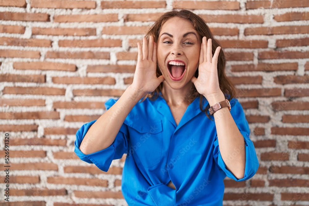 Poster beautiful brunette woman standing over bricks wall smiling cheerful playing peek a boo with hands sh