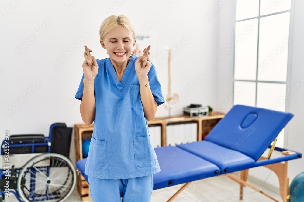 Sticker Beautiful caucasian physiotherapist woman working at pain recovery clinic gesturing finger crossed smiling with hope and eyes closed. luck and superstitious concept.