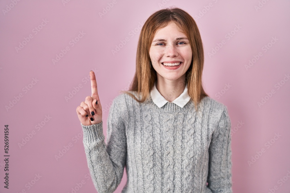 Poster beautiful woman standing over pink background showing and pointing up with finger number one while s