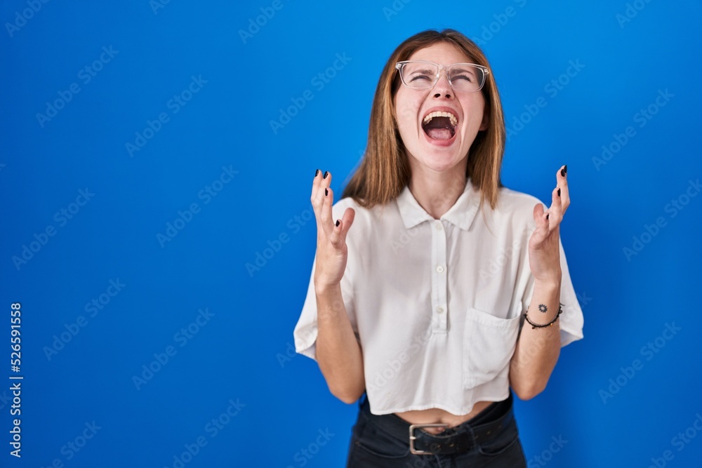 Sticker Beautiful woman standing over blue background crazy and mad shouting and yelling with aggressive expression and arms raised. frustration concept.