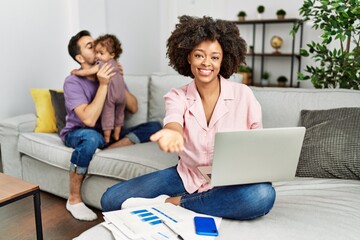 Mother of interracial family working using computer laptop at home smiling cheerful offering palm hand giving assistance and acceptance.