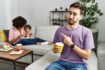 Hispanic father of interracial family drinking a cup coffee pointing aside worried and nervous with forefinger, concerned and surprised expression