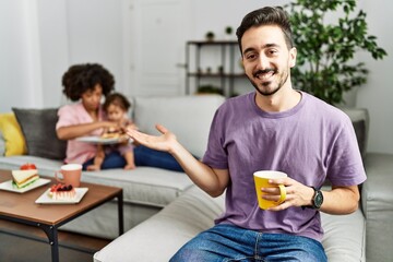 Hispanic father of interracial family drinking a cup coffee smiling cheerful presenting and pointing with palm of hand looking at the camera.