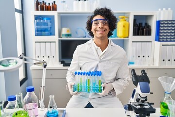 Hispanic man with curly hair working at scientist laboratory looking positive and happy standing and smiling with a confident smile showing teeth