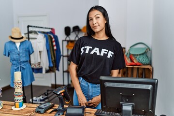 Young hispanic woman working as staff at retail boutique smiling looking to the side and staring away thinking.