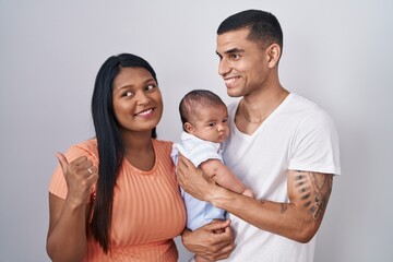 Young hispanic couple with baby standing together over isolated background smiling with happy face looking and pointing to the side with thumb up.