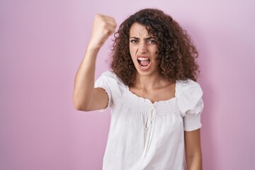 Hispanic woman with curly hair standing over pink background angry and mad raising fist frustrated...