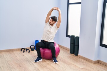 Young hispanic man sitting on fit ball stretching at sport center