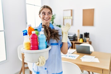 Young blonde woman wearing cleaner uniform holding cleaning products smiling cheerful presenting and pointing with palm of hand looking at the camera.