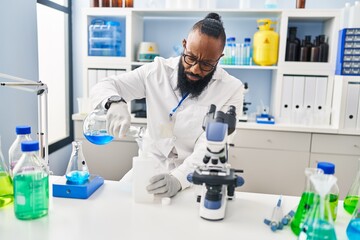 Naklejka premium Young african american man wearing scientist uniform measuring liquid at laboratory