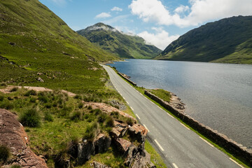 Small narrow road in a mountains by a lake with blue water with beautiful nature scenery. Warm sunny day, cloudy sky. Connemara area, Ireland. Irish landscape. Travel and sightseeing concept.