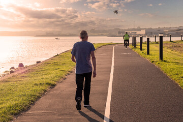 Man walking on a foot path in a park by the ocean at sunrise. Warm color tone. Cyclist out of focus...