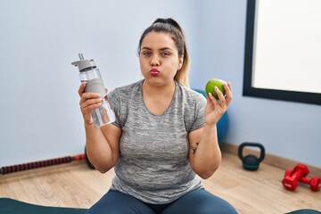 Young hispanic woman holding green apple and bottle of water puffing cheeks with funny face. mouth...
