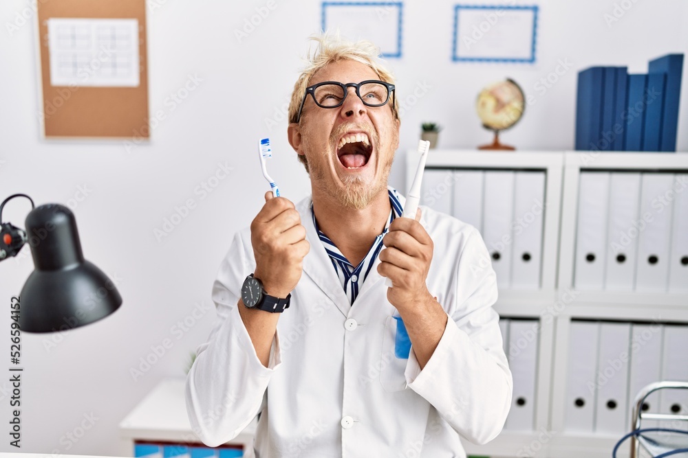 Sticker Young blond man working at dentist clinic holding electric toothbrush and teethbrush angry and mad screaming frustrated and furious, shouting with anger looking up.