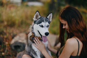 A woman with a husky breed dog smiles and affectionately strokes her beloved dog while walking in nature in the park in autumn against the backdrop of sunset, lifestyle in traveling with an animal