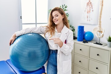 Young caucasian woman working at rehabilitation clinic holding pilates ball smiling happy pointing with hand and finger