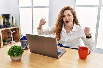 Young caucasian woman working at the office using computer laptop angry and mad raising fists frustrated and furious while shouting with anger. rage and aggressive concept.