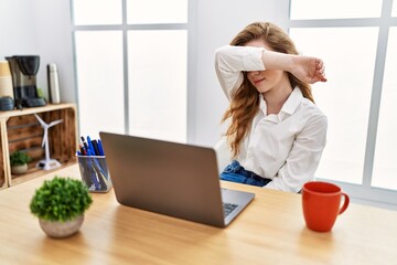 Young caucasian woman working at the office using computer laptop covering eyes with arm, looking serious and sad. sightless, hiding and rejection concept