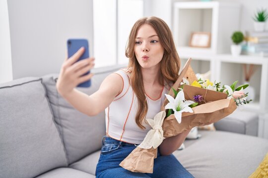 Caucasian woman holding bouquet of white flowers taking a selfie picture making fish face with mouth and squinting eyes, crazy and comical.