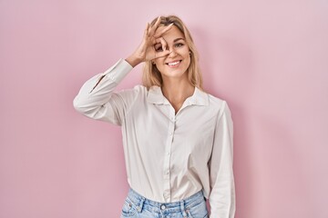 Young caucasian woman wearing casual white shirt over pink background doing ok gesture with hand smiling, eye looking through fingers with happy face.