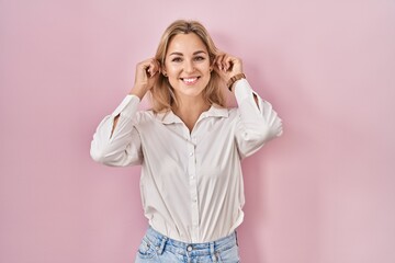 Young caucasian woman wearing casual white shirt over pink background smiling pulling ears with...