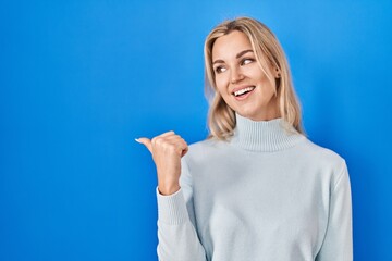 Young caucasian woman standing over blue background smiling with happy face looking and pointing to the side with thumb up.