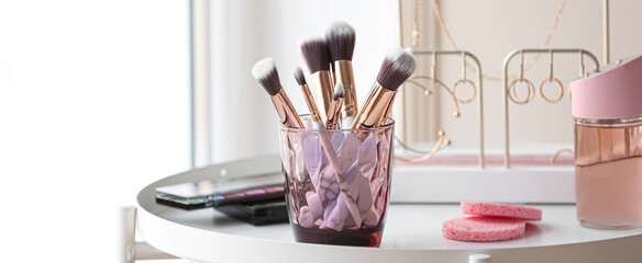 Holder with makeup brushes, cosmetics and sponges on table near window