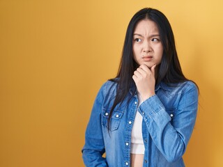 Young asian woman standing over yellow background thinking worried about a question, concerned and...