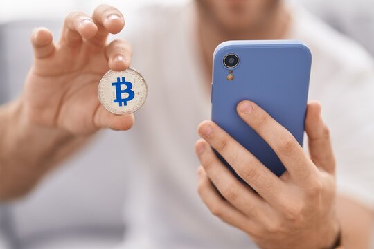 Young caucasian man using smartphone holding bitcoin at home